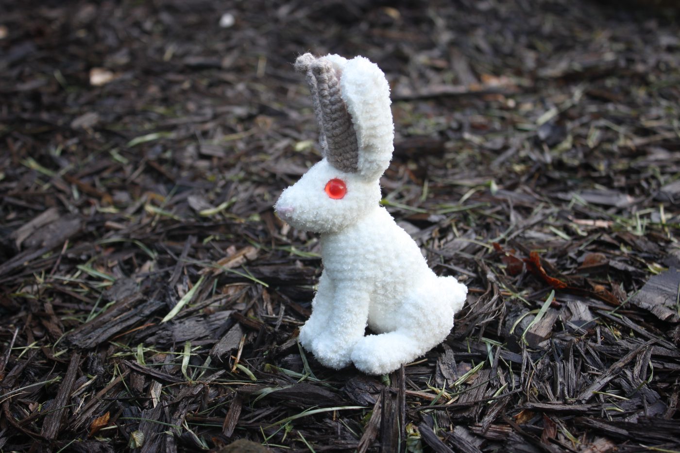 A small white crochet jackalope with a red gem eye, sitting on mulch