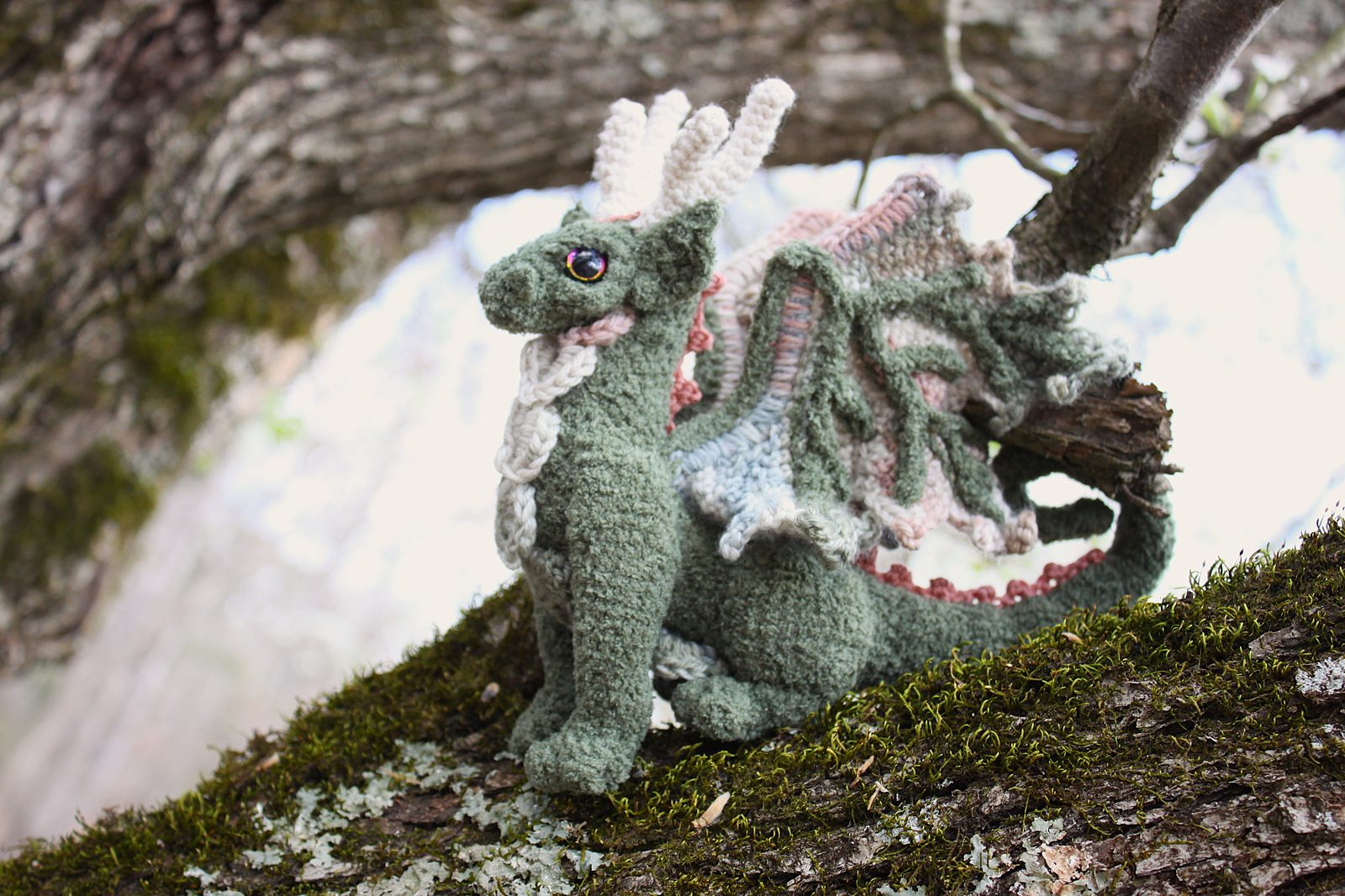 A moss-green crochet dragon with pink-and-cream wings, perched on a mossy tree branch