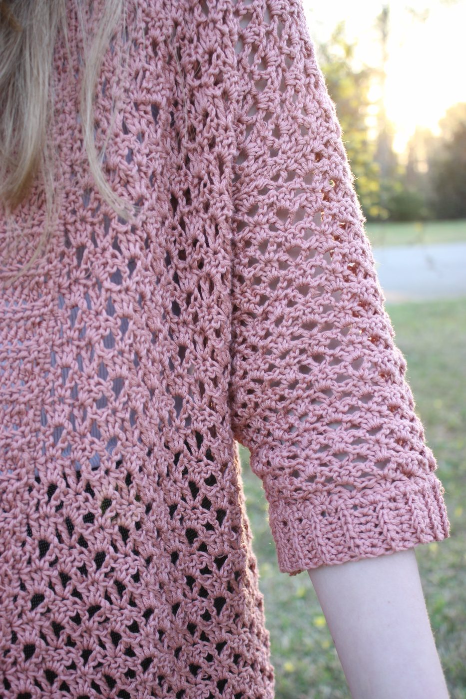 A dusty rose crochet sweater in an open-lace stitch, outdoors at golden hour